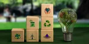 Bloques de madera con íconos de reciclaje, energía y sostenibilidad junto a un foco con una planta verde, representando cómo las empresas integran el valor compartido.